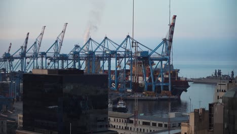 Valparaiso-buildings-in-front,-container-cargo-ship-docked-waiting-to-be-loaded-near-cranes-in-Sea-Port,-Chile