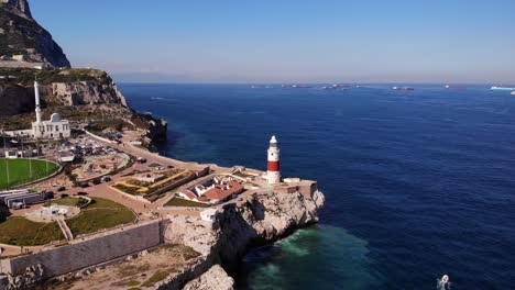 Rot-weiß-Gestreifter-Leuchtturm-Und-Eine-Große-Moschee-Am-Europa-Point-In-Gibraltar-Mit-Blick-Auf-Mehrere-Frachter-Vor-Anker,-Während-Die-Mittelmeerdünung-Gegen-Die-Klippen-Spritzt
