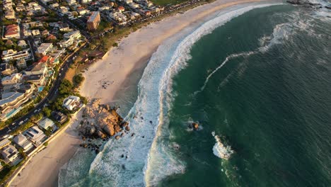 beautiful-white-sand-beach-at-Camps-Bay,-Cape-Town-with-turquoise-ocean-waves-crashing-at-sunset,-aerial
