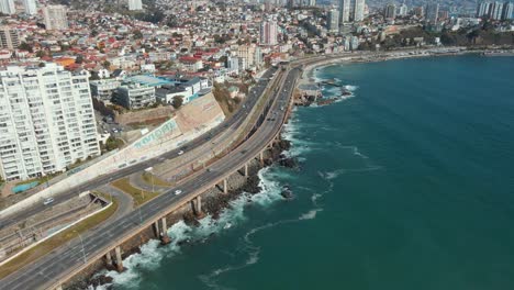 Aerial-dolly-in-of-traffic-in-avenue-next-to-turquoise-sea-coastine,-Viña-del-Mar-city-buildings-and-hills-in-back,-Chile