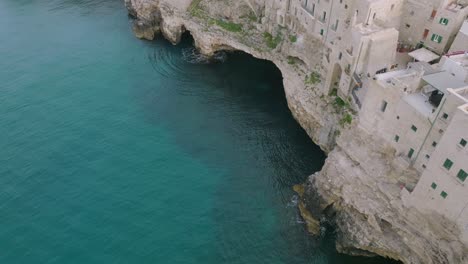Aerial-pan-up-footage-of-blue-sea-crashing-against-the-cliffs-in-Polignano-a-Mare,-Italy