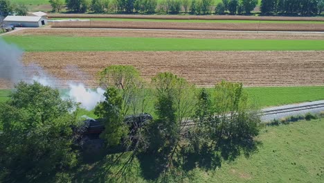A-Drone-Side-View-of-an-Antique-Steam-Passenger-Train-in-Slow-Motion-Blowing-Smoke-and-Steam-Traveling-Thru-Fertile-Corn-Fields-and-Traveling-Through-Trees-on-a-Sunny-Summer-Day