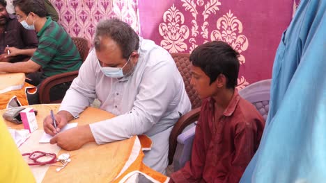 Un-Médico-Con-Mascarilla-Y-Recetando-Medicamentos-A-La-Víctima-De-La-Inundación-En-La-Carpa-Instalada-Para-El-Campamento-De-Ayuda-Contra-Inundaciones-En-Sindh,-Pakistán