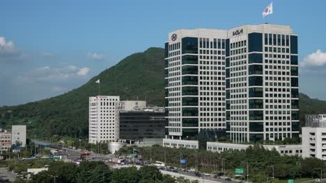 Hyundai-Kia-Corporate-Buildings-with-Seoul-skyline,-car-traffic-on-Yangjae-daero-against-Guryongsan-mountain-in-summer