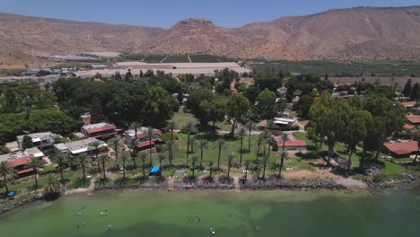 A-drone-flies-back-and-shows-Kibbutz-Ein-Gev-Beach---Israel,-Sea-of-Galilee,-The-Golan-Heights-in-the-background-and-people-inside-the-Sea-of-Galilee
