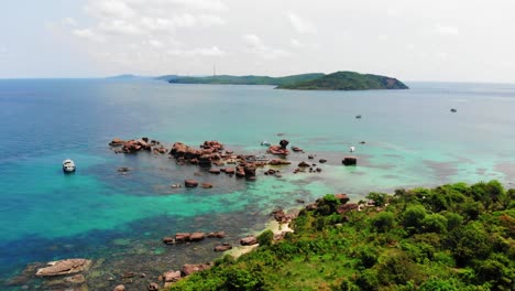 Boat-Anchored-in-Clear-Turquoise-Sea-Waters-next-to-Rocks-and-Rocky-Coast-of-Gam-Ghi-island-in-Vietnam-Archipel,-Phu-Quoc-Region,-Aerial-View