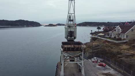 Old-rusty-harbor-crane-in-Lillesand-Norway---Aerial-rotating-around-crane-wit-grab-and-north-sea-in-background