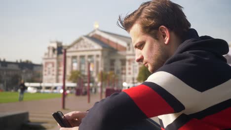 Handsome-young-man-typing-on-mobile-near-the-Concert-Hall-in-Amsterdam-in-a-a-sunny-day