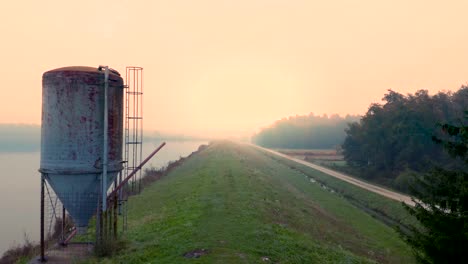 Landscape-with-a-lake-and-silos-for-fish-feeding-and-a-single-road-bordering-it-at-misty-autumn-morning-in-Central-Europe