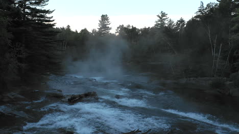 Aerial-drone-shot-flying-upstream-over-a-dark-misty-forest-river-waterfall-at-sunset-with-trees-in-silhouette