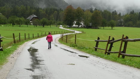 Little-girl-playfully-walking-in-rain-on-country-road,-farmhouse-in-background,-alpine-valley,-zoom-in,-from-behind-facing-away