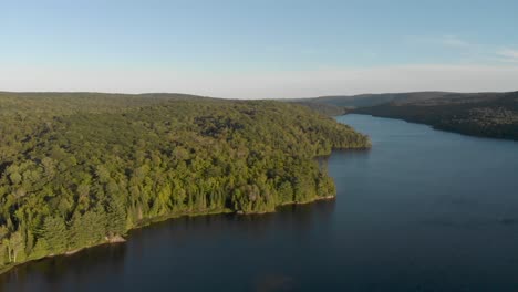 Slow-aerial-view-of-lake-in-quebec-surrounded-by-trees-at-sunrise-with-the-sun-shining-over-the-hill-and-trees