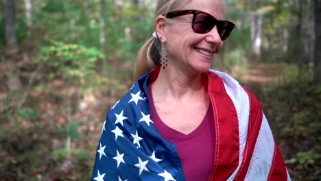 Camera-turning-around-a-pretty,-blonde-woman-in-a-forest-with-an-American-flag-wrapped-around-her