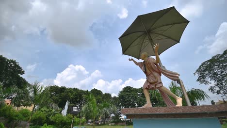 Ein-Zeitraffer-Der-Statue-Des-Indischen-Halbgottes-Chaitanya-Deb-Mit-Regenschirm-Und-Wolken-Am-Blauen-Himmel-In-Der-Nähe-Eines-Hindu-tempels