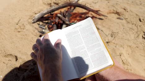 a-close-shot-slow-motion-shot-of-a-man-opening-up-a-bible-besides-a-fire-on-the-beach