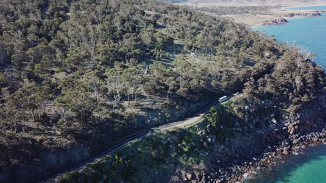 Drone-Pan-Down-Hill-With-Cars-Driving-Down-Stunning-Road-Near-Coast-With-Trees-And-Blue-Ocean-In-Tasmania-Australia