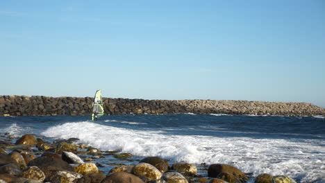 Pan-shot-of-a-norwegian-windsurfer,-surfing-a-wave-close-to-the-rocky-shore,-on-the-North-sea,-in-the-atlantic-ocean,-on-a-sunny,-summer-day,-in-Lista,-South-Norway