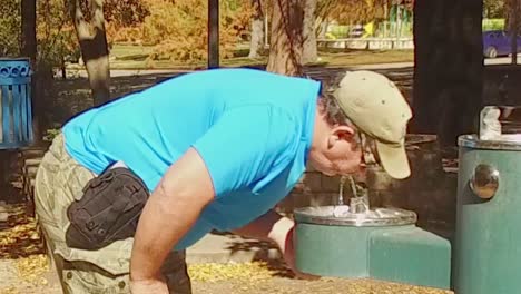 Man-drinking-from-drinking-fountain-in-the-park-on-a-beautiful-sunny-day-surrounded-by-trees-and-grass