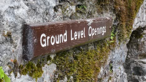 Brown-Wooden-Ground-Level-Openings-Sign-on-Mossy-Rock-Wall-Close-Up