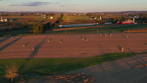 Amazing-aerial-over-farmland-and-bronze-coloured-pasture-dotted-with-bales-of-hay-during-sunset