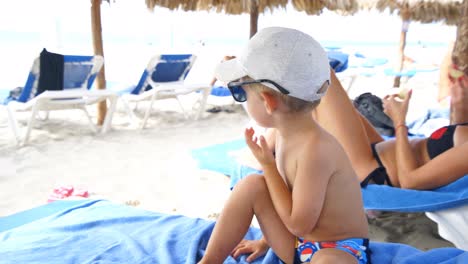 Child-sitting-on-the-sunbed-at-the-beach-plays-with-his-hat-and-sunglasses-removing-them-and-putting-them-several-times