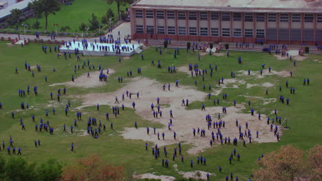 School-students-playing-in-the-ground-aerial-zoom-out-of-the-ground,-some-running-and-having-fun,-students-are-in-blue-pants-and-shirts,-school-building-in-the-background