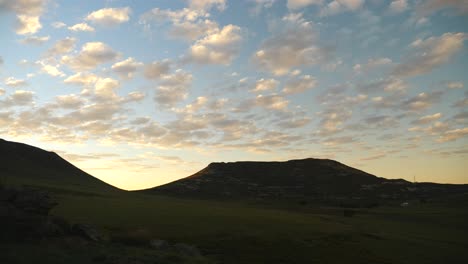 Timelapse:-Early-morning,-small,-softly-lit-clouds-travel-across-the-sky-before-sunrise,-with-grassy-landscape-and-sandstone-hills-slowly-lighting-up-and-turning-green-on-a-summer-morning-in-Lesotho