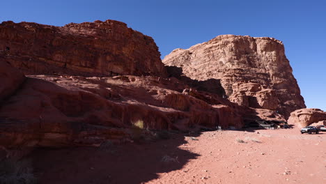 Enormes-Montañas-De-Roca-Roja-En-Medio-Del-Desierto-De-Wadi-Rum-En-Un-Día-Soleado-Con-Turistas-Escalando-La-Colina-Y-Beduinos-Jeps-Esperando