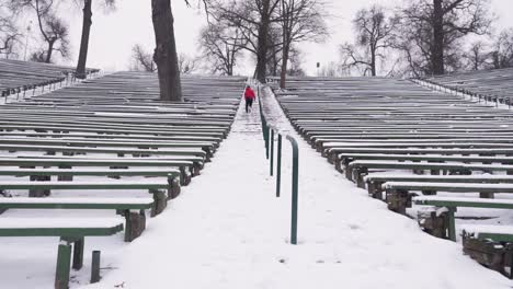 Young-man-with-red-hoodie-and-black-pants-is-running-up-stairs-outside-in-the-amphitheater-in-winter