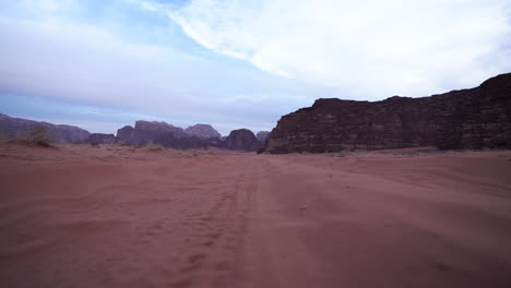 Niedrigwinkelaufnahme-Der-Kamera,-Die-Sich-Auf-Der-Sandstraße-In-Der-Wadi-Rum-Wüste-Mit-Wolken-Und-Blauem-Himmel-Im-Hintergrund-Bewegt