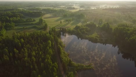 Misty-morning-sunlight-over-autumn-forest-lake-and-scenic-countryside-landscape-aerial-wide-view