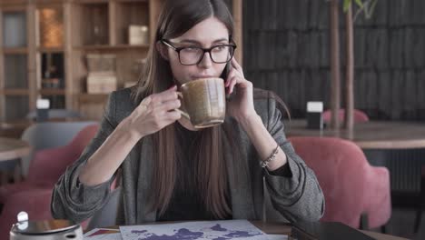 Beautiful-young-business-lady-in-a-glasses-is-sitting-in-a-cafe,-drinking-a-coffee,-talking-on-a-phone-and-studying-some-diagrams-and-statistics