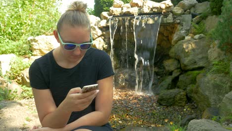 Sexy-Woman-In-Shades-Browses-Social-Media-On-Her-Smartphone-While-Sitting-In-A-Park-Next-To-A-Waterfall