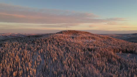 Aerial-view-of-sunset-over-snowy-frozen-hill-in-deep-forest-with-mountains