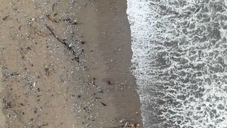 Slow-motion-aerial-shot-as-waves-washing-out-garbage-from-the-sea,-polluted-ocean-coast