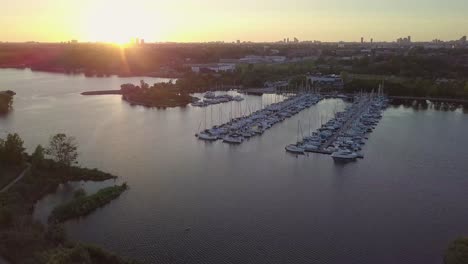 Aerial-Sunset-Wide-Shot-Flying-Toward-Sailboat-Marina-Yacht-Club-Dock-In-Lake-Bay-Among-Green-Trees-And-Close-Birds-With-City-Buildings-Skyline-In-Background-In-Toronto-Ontario-Canada