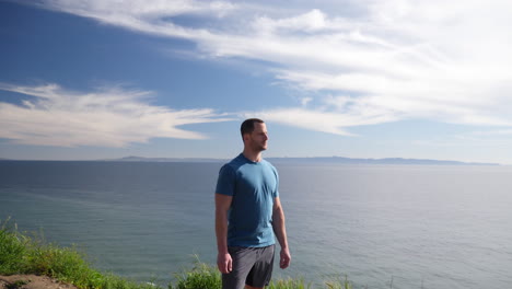 A-fit-strong-man-standing-proud-on-the-edge-of-an-ocean-cliff-nature-hiking-trail-with-an-epic-beach-view-on-a-sunny-day-in-Santa-Barbara,-California-SLOW-MOTION