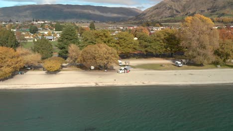 Aerial-Drone-ascending-from-the-beach-overviewing-the-town-and-Lake-Wanaka,-New-Zealand-in-autumn