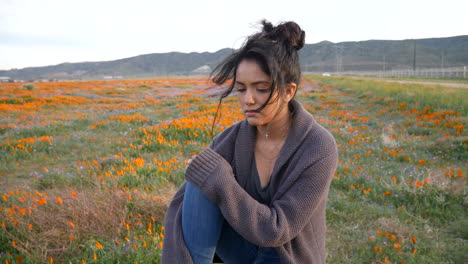 A-young-pretty-girl-sitting-on-a-rock-in-a-field-of-wild-flowers-looking-sad-and-lonely-with-black-hair-blowing-in-the-wind-in-slow-motion