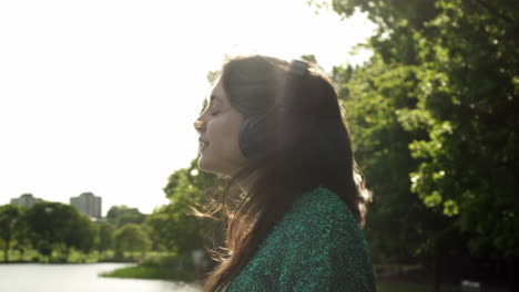 Attractive-young-Italian-woman-with-headphones-listening-to-music-at-the-park-with-a-sunset-look-around-smile,-dancing,-enjoying-life