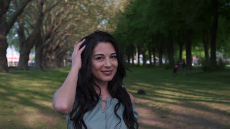 Attractive-and-playful-latina-woman-with-black-wavy-hair-walking-under-the-trees-in-a-park-in-London,-looking-at-the-camera,-happy-with-a-beautiful-smile