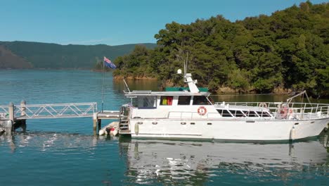 SLOWMO---Cruiser-and-small-boat-docked-at-in-bay-in-Marlborough-Sounds,-New-Zealand---Aerial-fyling-low-side-shot