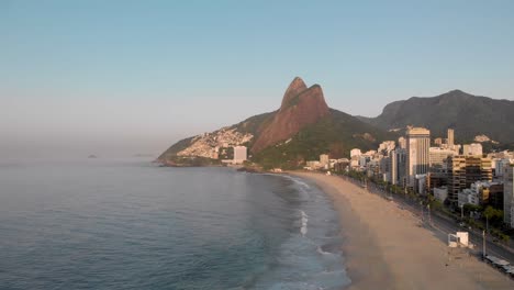 Forwards-and-upwards-aerial-view-of-deserted-coastal-city-beach-of-Rio-de-Janeiro-during-early-morning-golden-hour