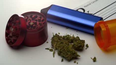 A-small-pile-of-ground-marijuana-lies-on-table-surrounded-by-a-grinder,-vaporizer,-open-pill-bottle-and-prescription-note