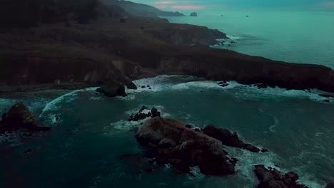 Fast-dolly-out-reveal-of-huge-ocean-cliff-peninsula-eroded-by-crashing-waves-at-dusk-at-Sand-Dollar-Beach-in-Big-Sur-California