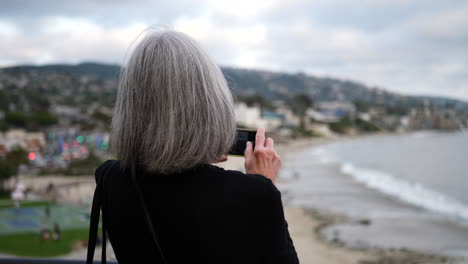 Una-Anciana-De-Vacaciones-Tomando-Una-Foto-Con-Su-Teléfono-De-La-Ciudad-Y-El-Océano-En-Laguna-Beach,-California-A-Cámara-Lenta