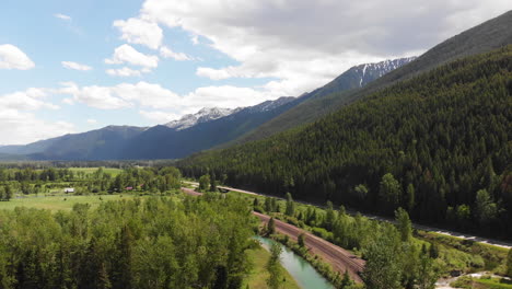 Aerial-shot-of-road,-river,-and-train-tracks-winding-through-East-Glacier-Park,-Montana,-snow-capped-mountains-and-forested-hills-in-the-background