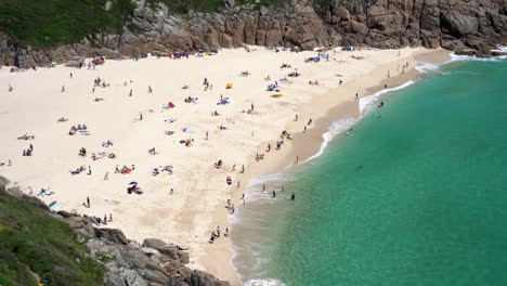 Aerial-view-of-Porthcurno-Beach-in-Cornwall-full-of-tourists,-swimmers,-surfers-and-families-having-fun-on-a-beautiful-spring-sunny-day