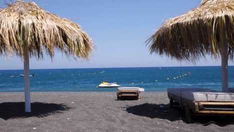 Handheld-Moving-Wide-Shot-of-Perissa-Beach-in-Santorini-Greece,-With-Black-Lava-Sand-on-a-Windy-and-Sunny-Summer-Day
