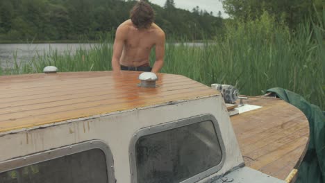 Young-topless-carpenter-walking-to-bow-of-wooden-boat-to-start-maintenance